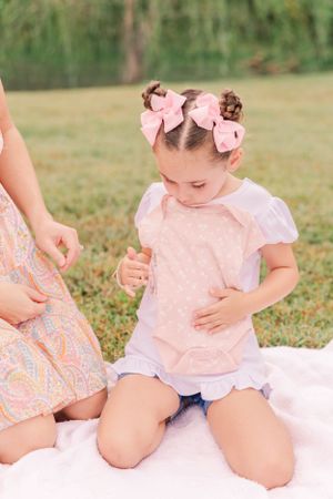 Baby wearing pink outfit and bows sits on white blanket in grassy outdoor setting.