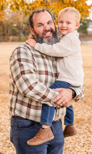 Adult in plaid shirt holding young child during fall outdoor portrait session.