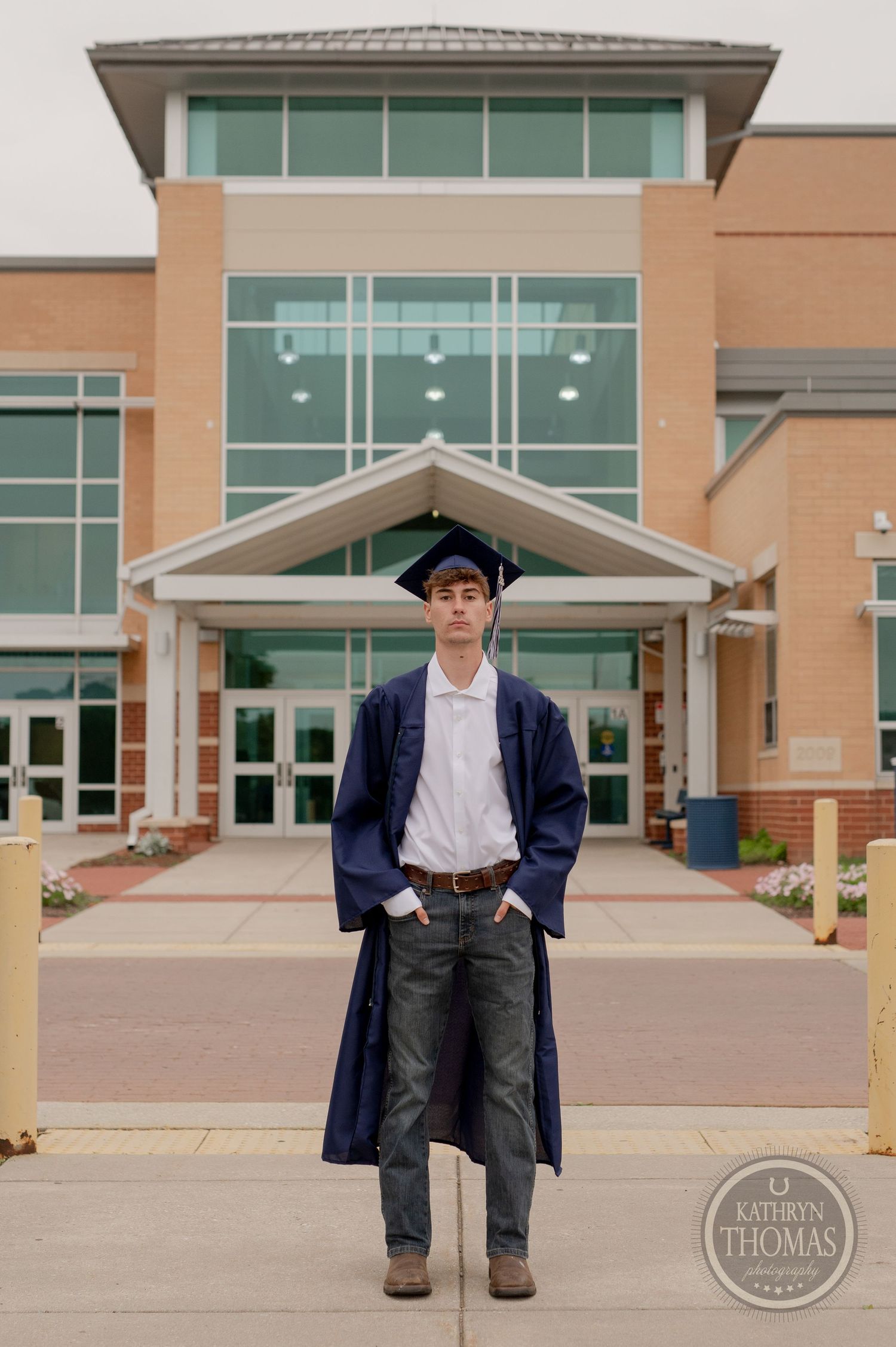 Graduate poses in front of school building wearing graduation regalia and casual attire.