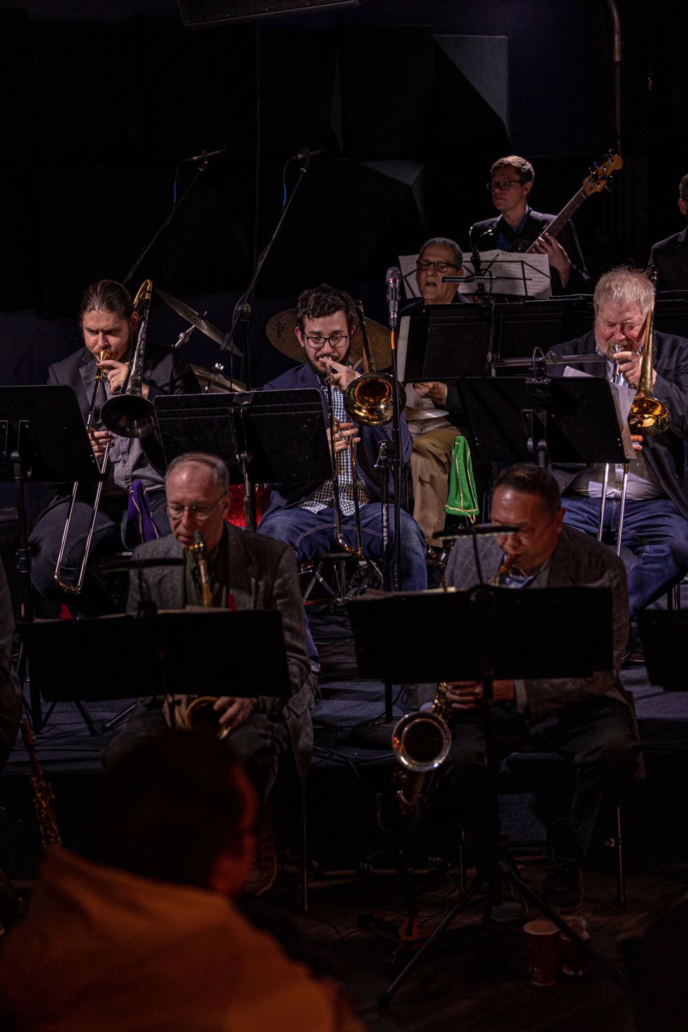 Jazz musicians perform on stage with brass instruments and music stands during a dimly lit concert performance.