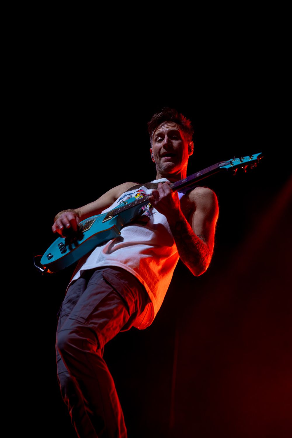 Guitarist performs energetically under red stage lighting against dark background.