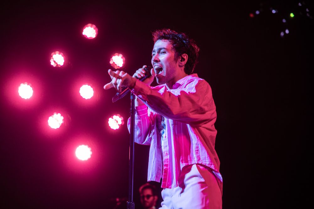 Concert performer singing energetically against a backdrop of pink stage lighting and glowing orbs.