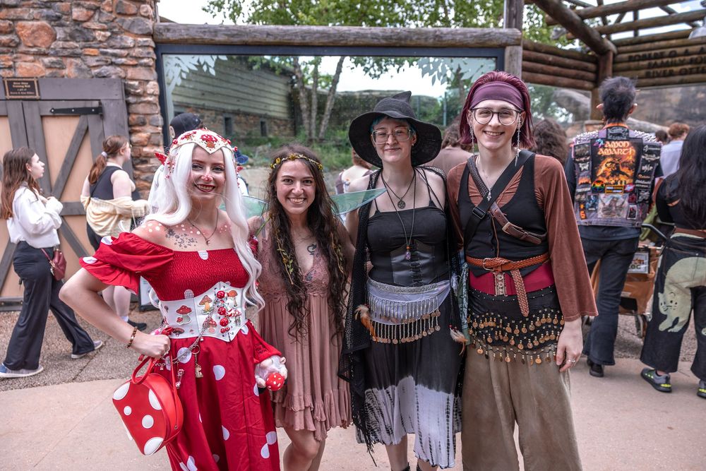 A group in costumes including a polka dot dress, western wear, and pirate-themed outfits pose together at an outdoor event.