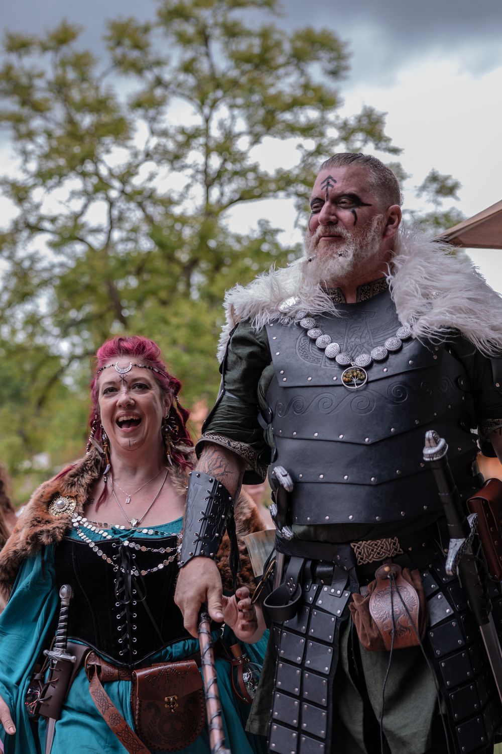 A pair of medieval faire participants laugh together while wearing period costumes with armor and decorative garments.