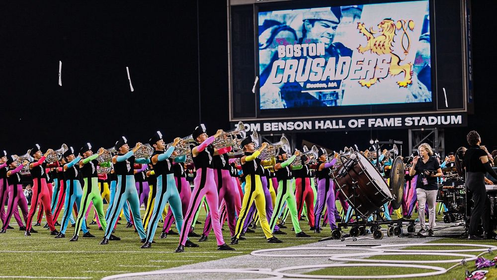 Boston Crusaders in vibrant neon costumes stand in synchronized formation on a football field at night.