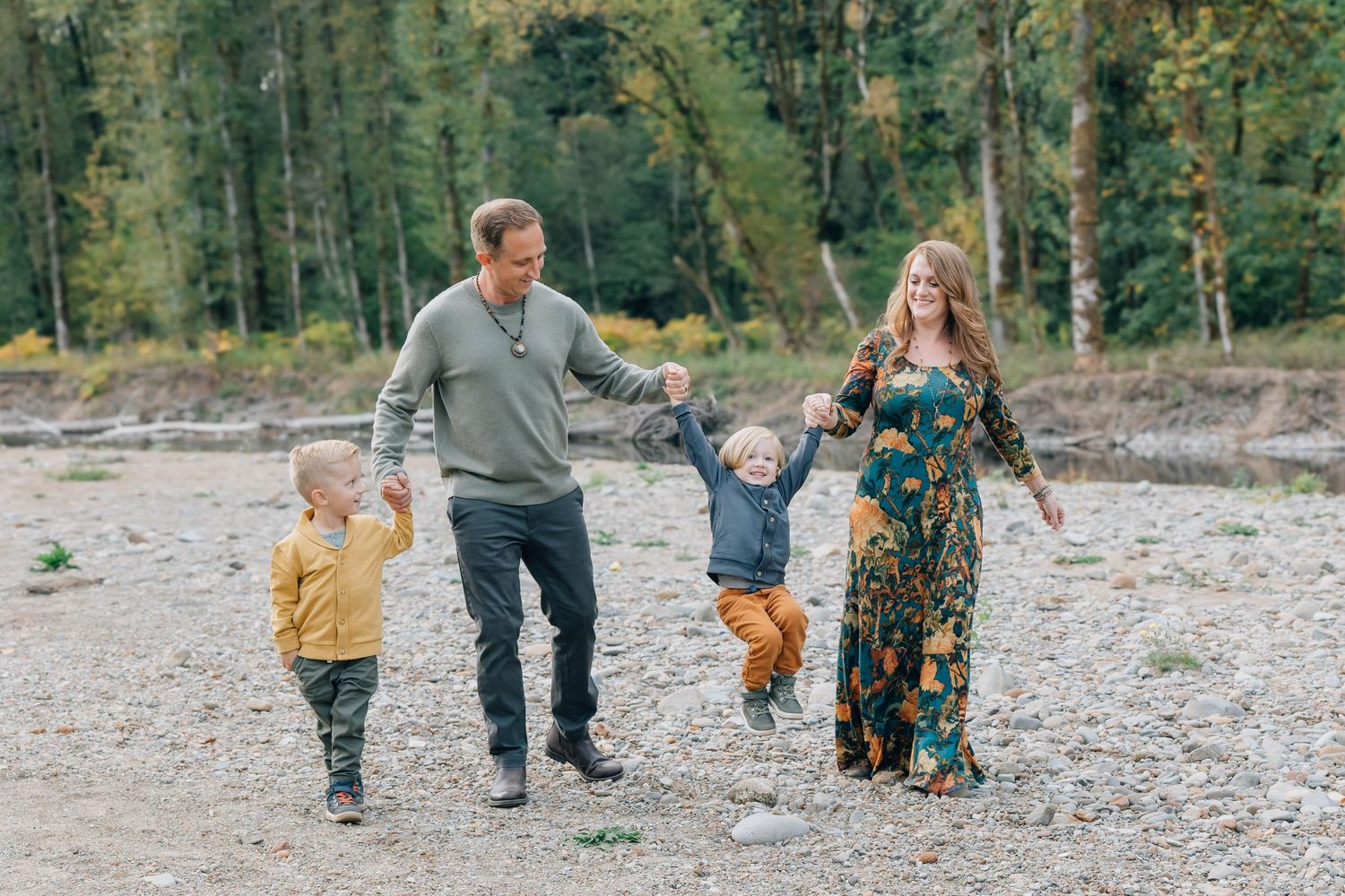 Joyful family of four swinging young boy between parents on a rocky riverbank outdoors.