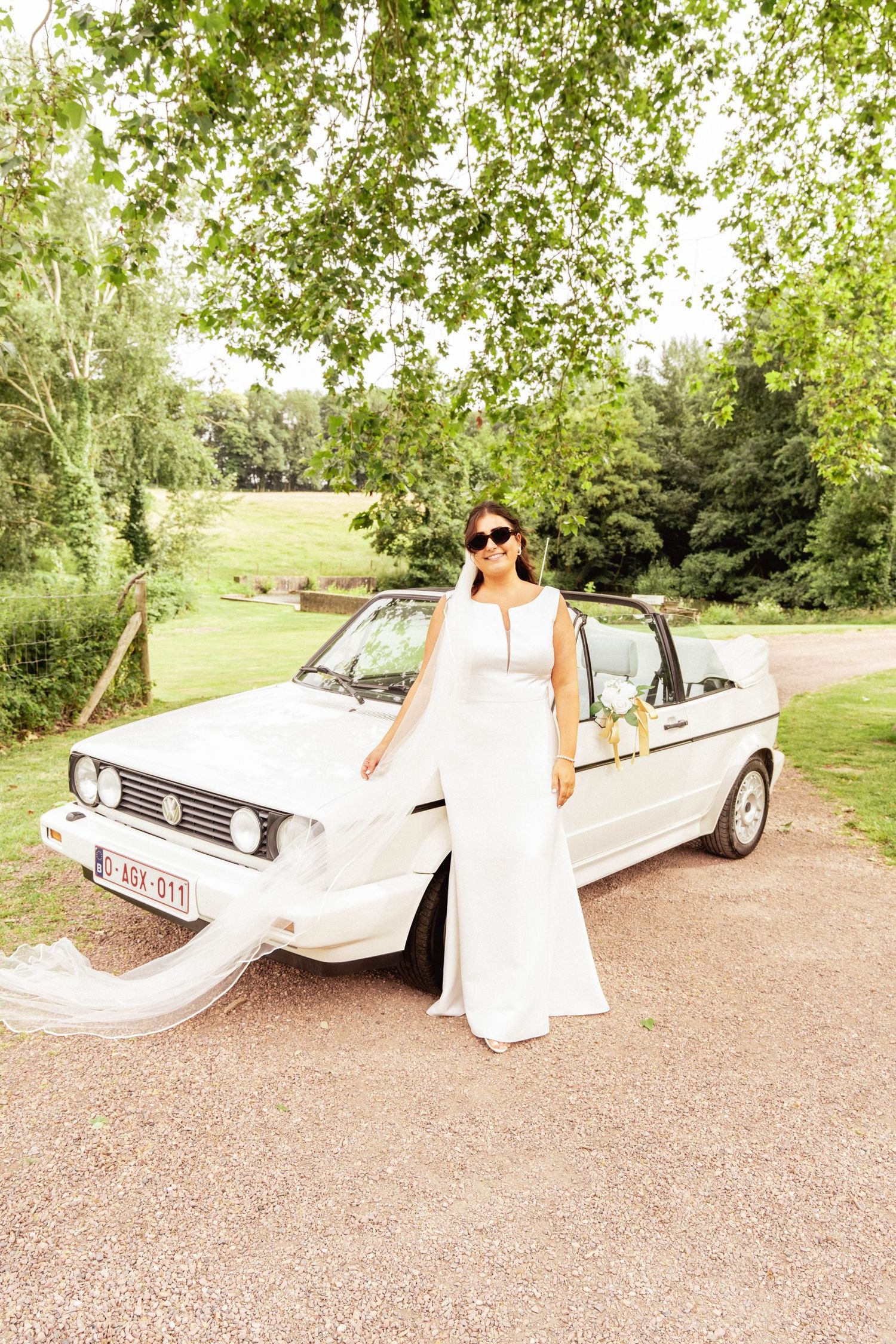 A classic white Volkswagen Golf convertible serves as an elegant wedding car on a tree-lined path.