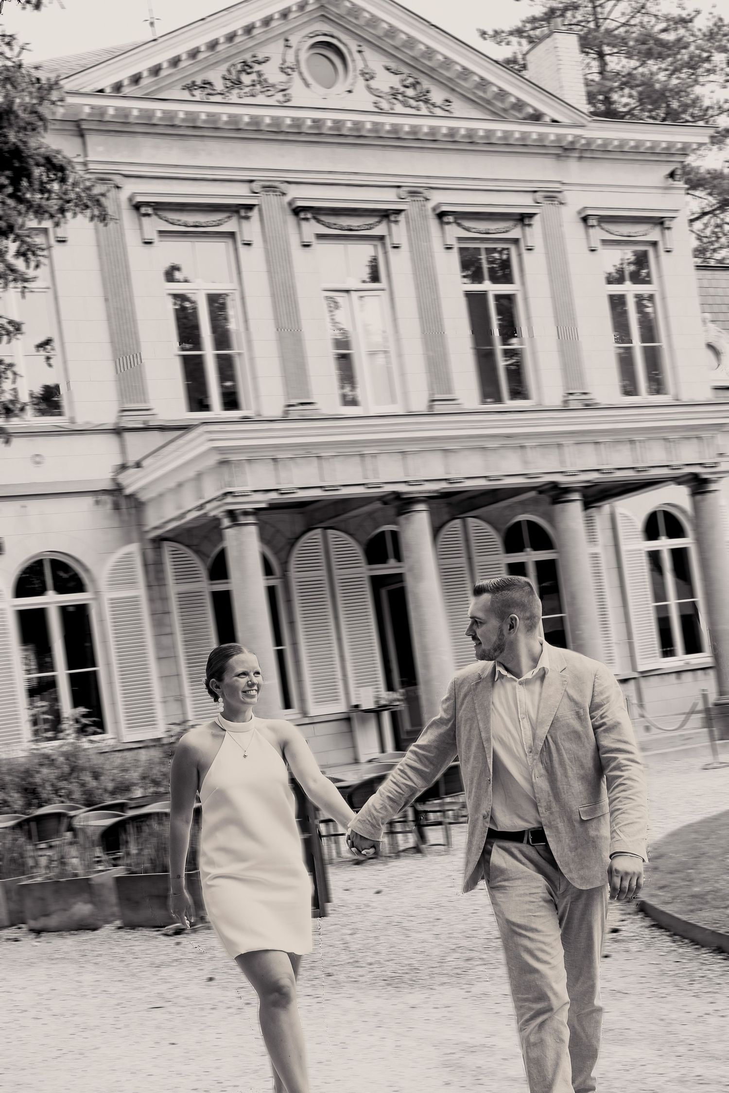 A couple strolls hand in hand outside a grand neoclassical building with white columns and arched windows.