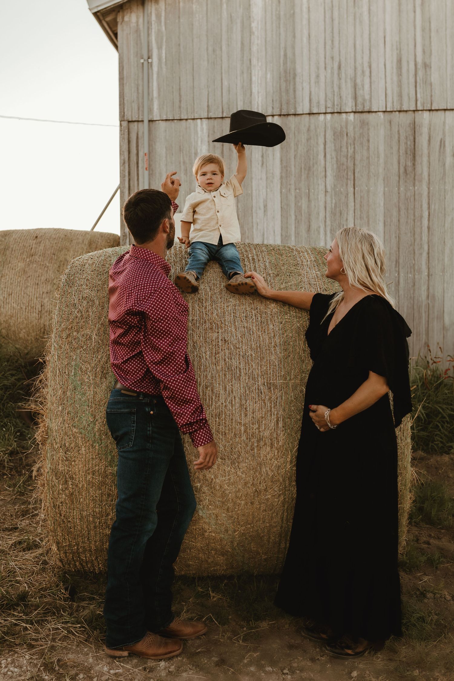 Family posing playfully with child on hay bales near rustic barn in countryside setting.