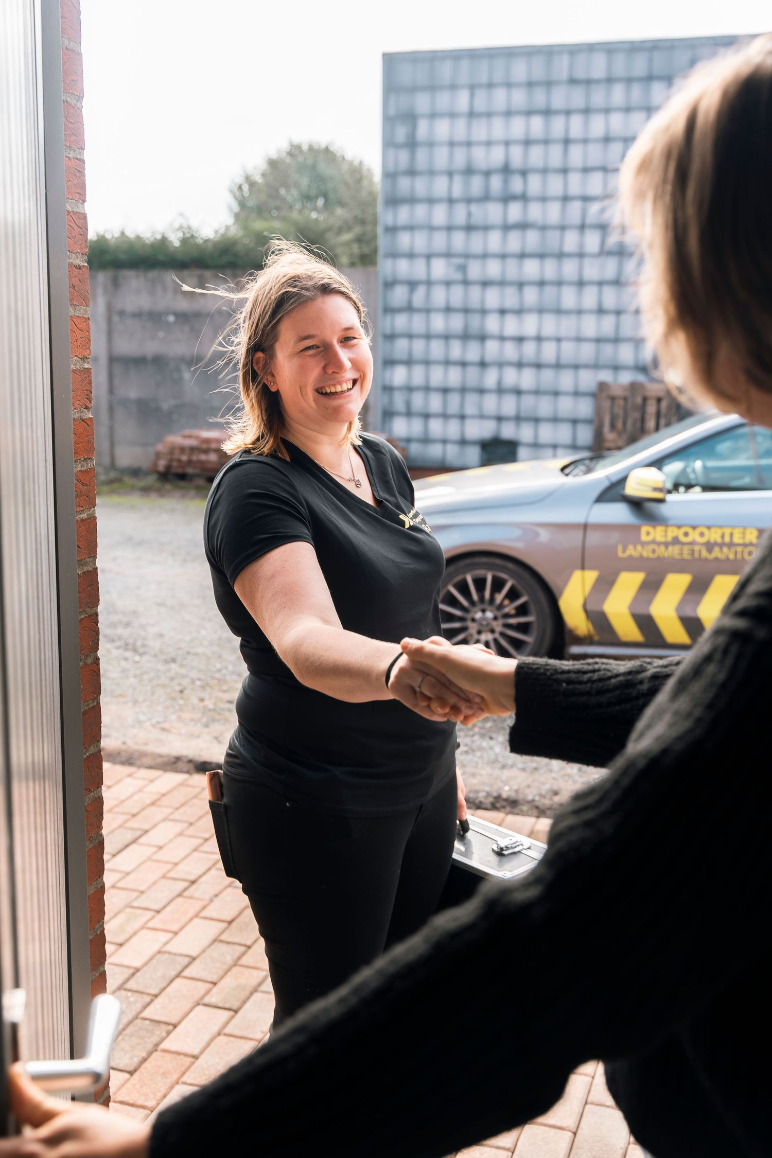 A person in black attire greets someone at a doorway with a delivery vehicle visible in the background.
