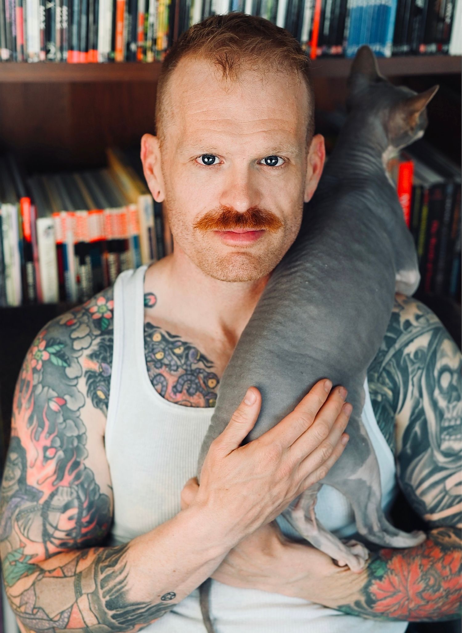 A person with colorful tattoos poses intimately with a grey pillow in front of a bookshelf.