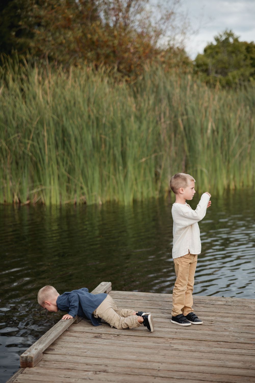 Two young children play on a wooden dock by a lake surrounded by tall reeds and autumn foliage.