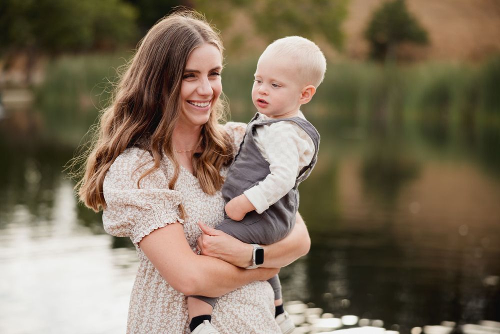 A mother in a floral dress holds a baby while standing on a dock by a peaceful lake.
