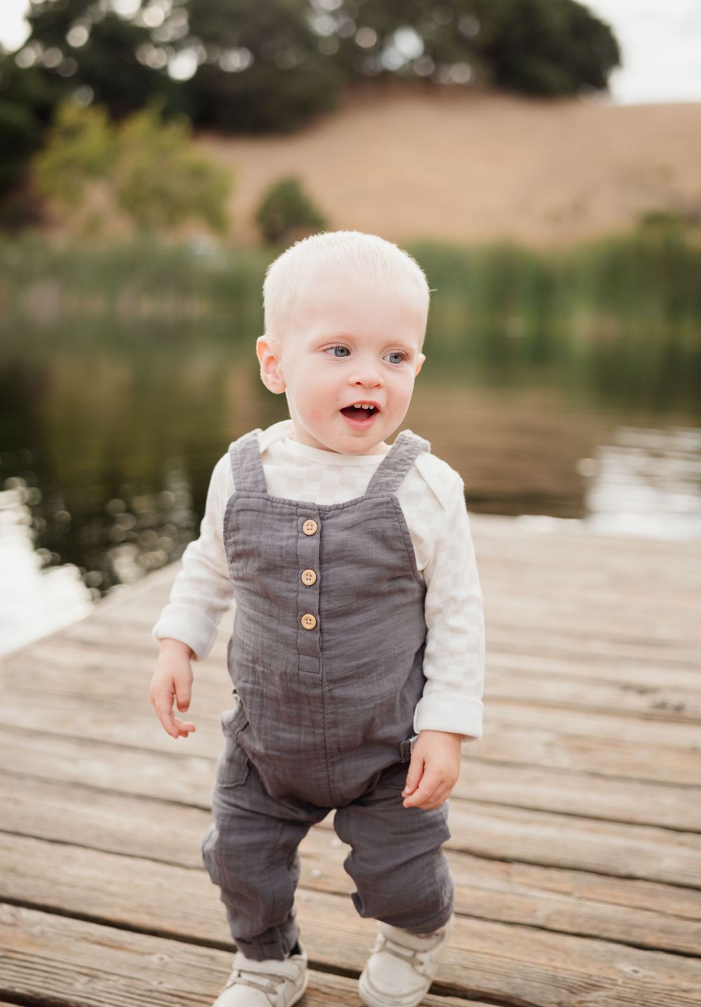 A baby in gray overalls stands smiling on a wooden dock with a lake in the background.