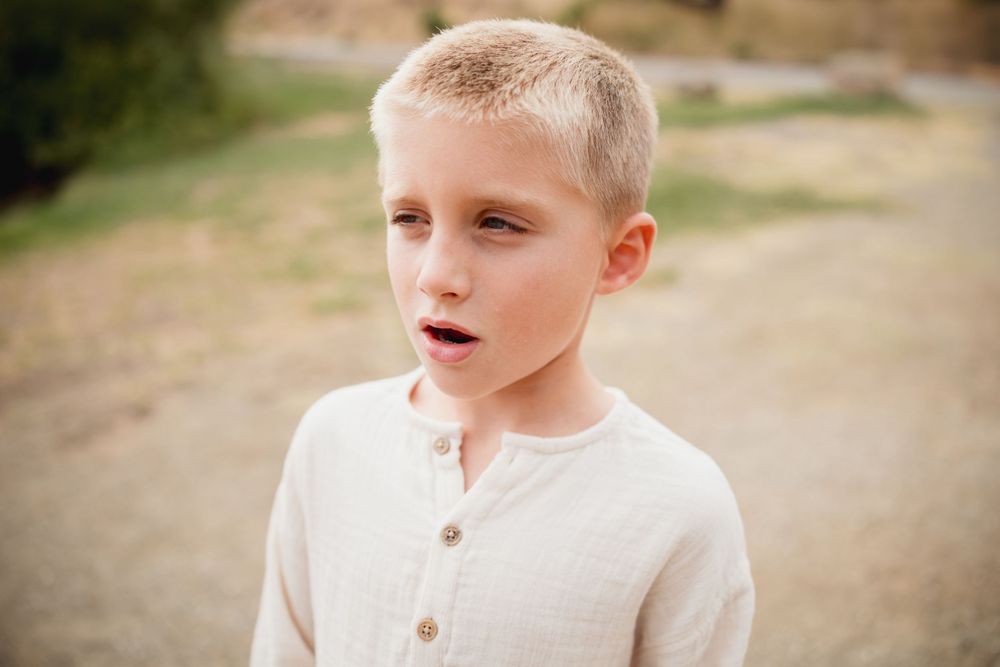 A child in a white henley shirt poses outdoors against a natural hillside background.