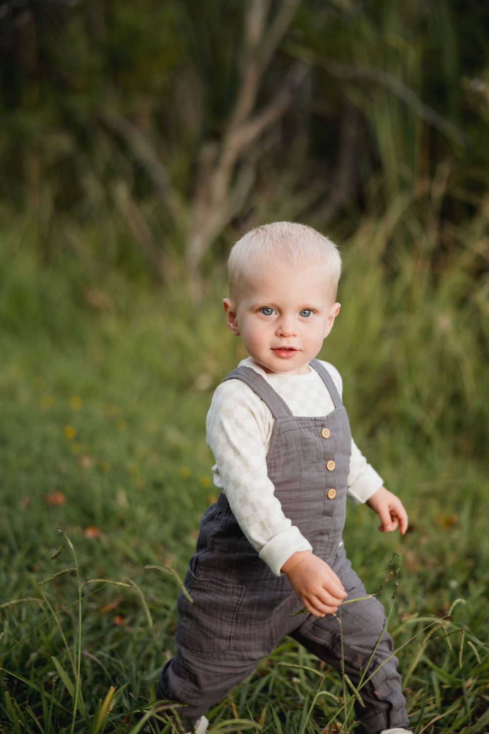 A toddler in gray overalls explores tall grass near a lake.