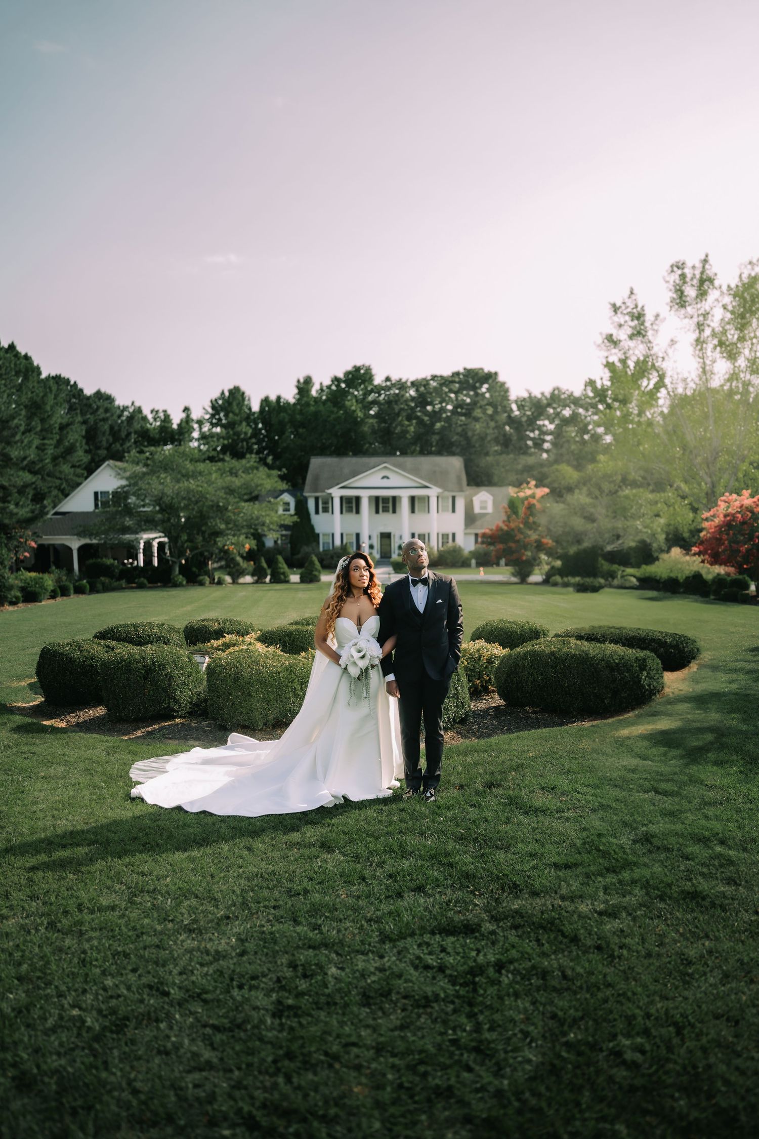 A bride and groom stand together on a manicured lawn with a white columned estate house in the background at sunset.