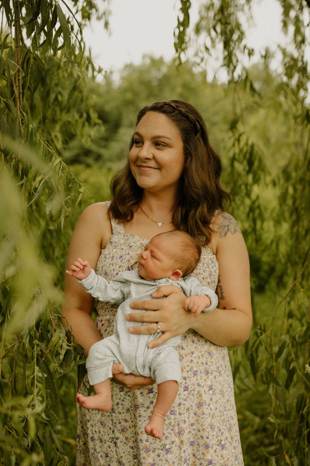 A parent holds a sleeping infant while standing under a willow tree in a floral dress.