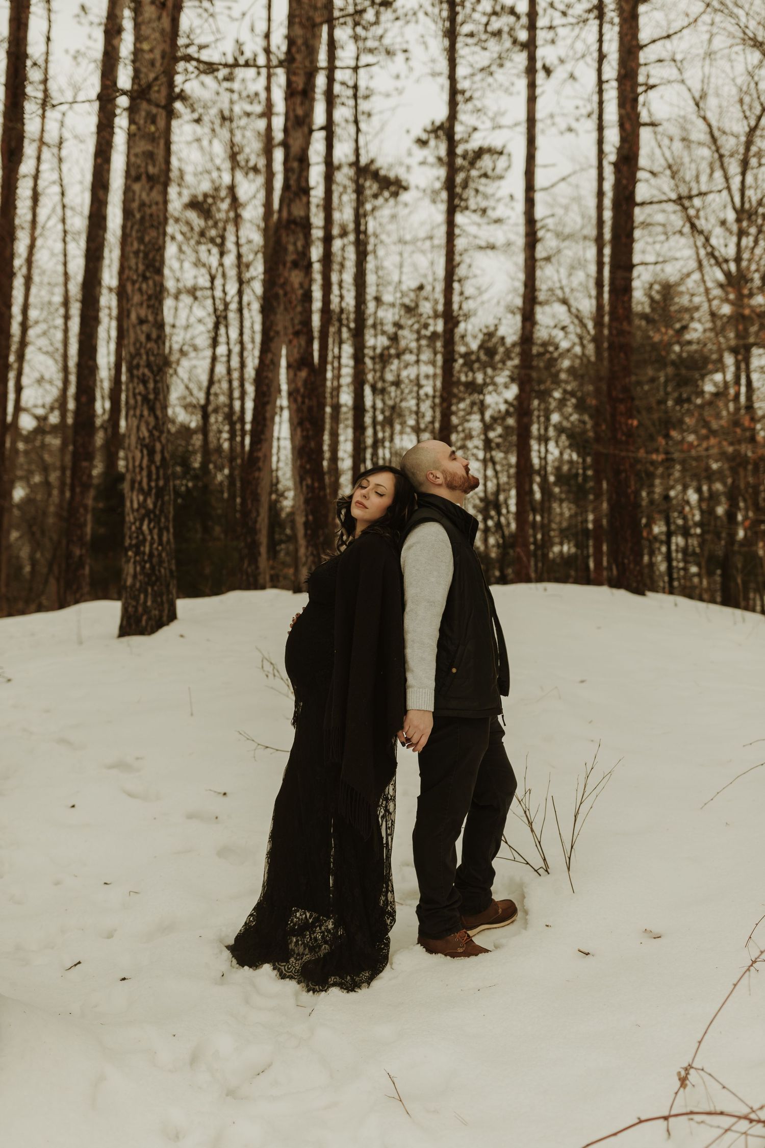 A couple shares a tender moment in winter snow among tall pine trees.