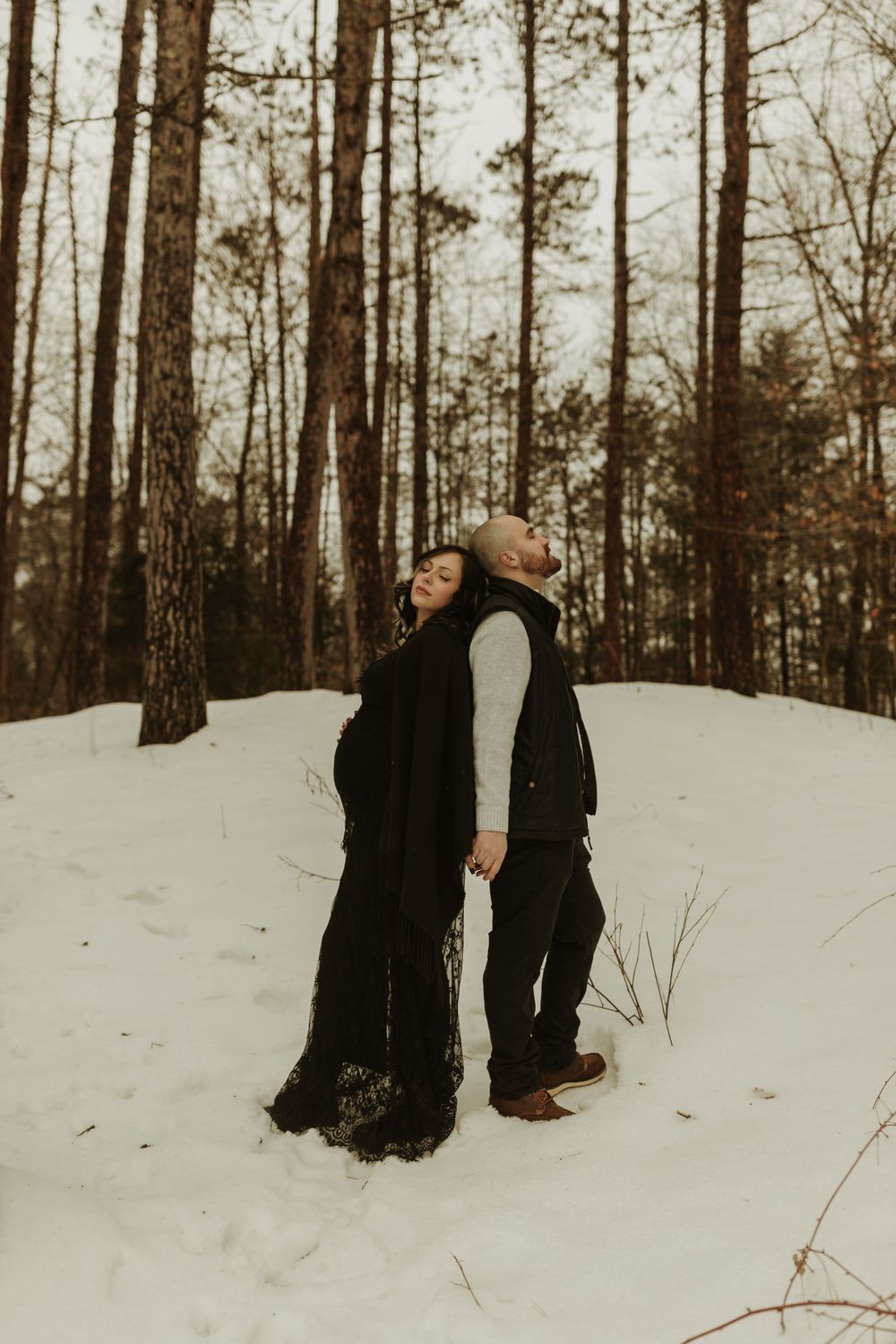 A couple shares a tender moment in winter snow among tall pine trees.