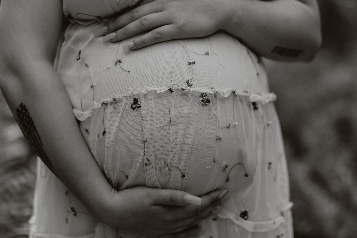 Close-up black and white photo of hands cradling a pregnant belly in a floral dress.