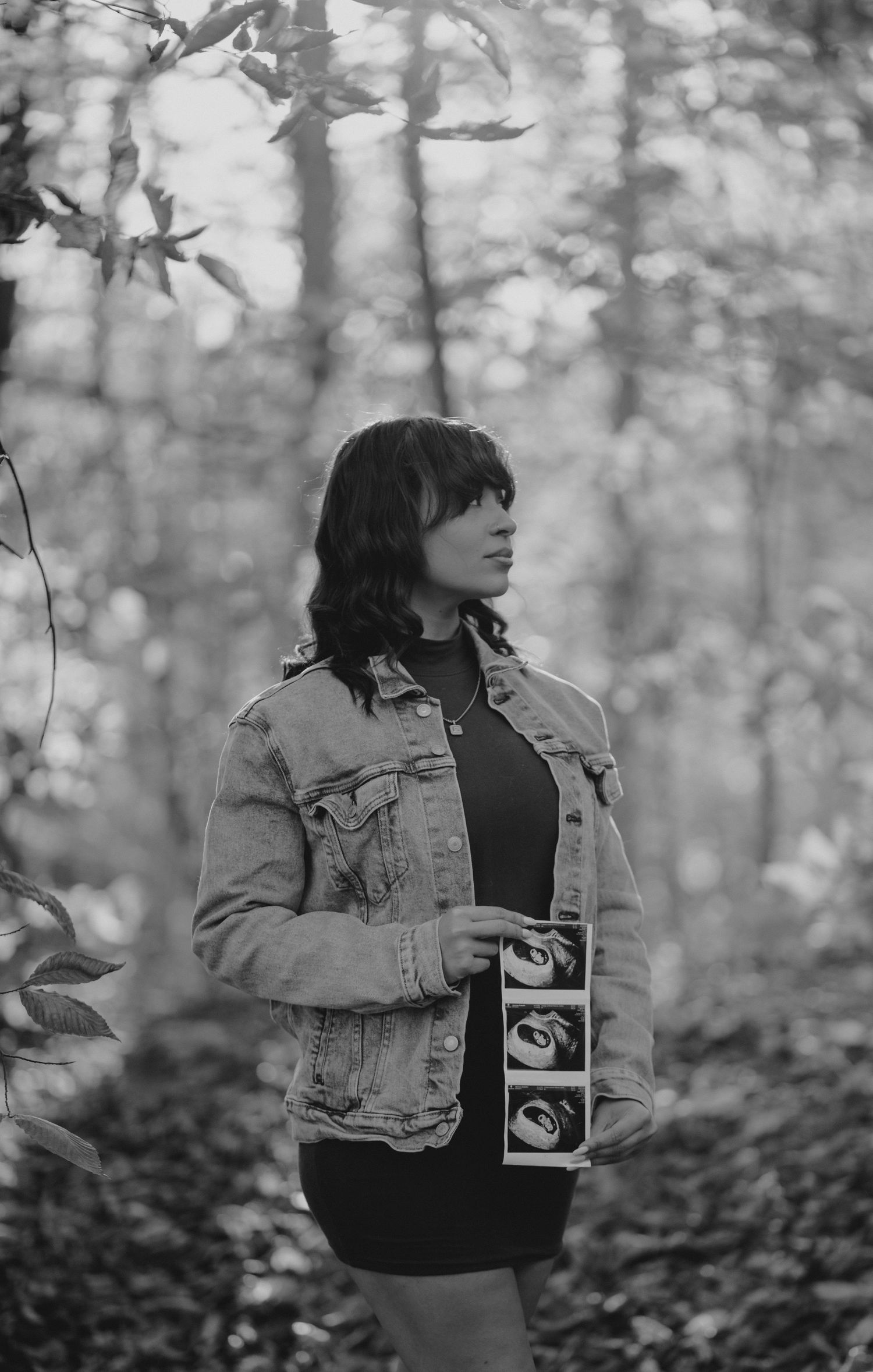 Black and white portrait in autumn woods holding ultrasound photos while wearing denim jacket.