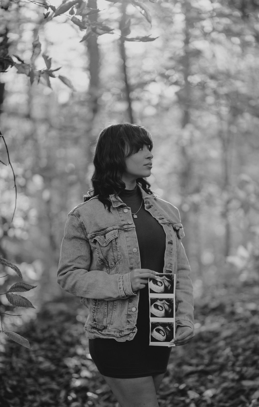 Black and white portrait in autumn woods holding ultrasound photos while wearing denim jacket.