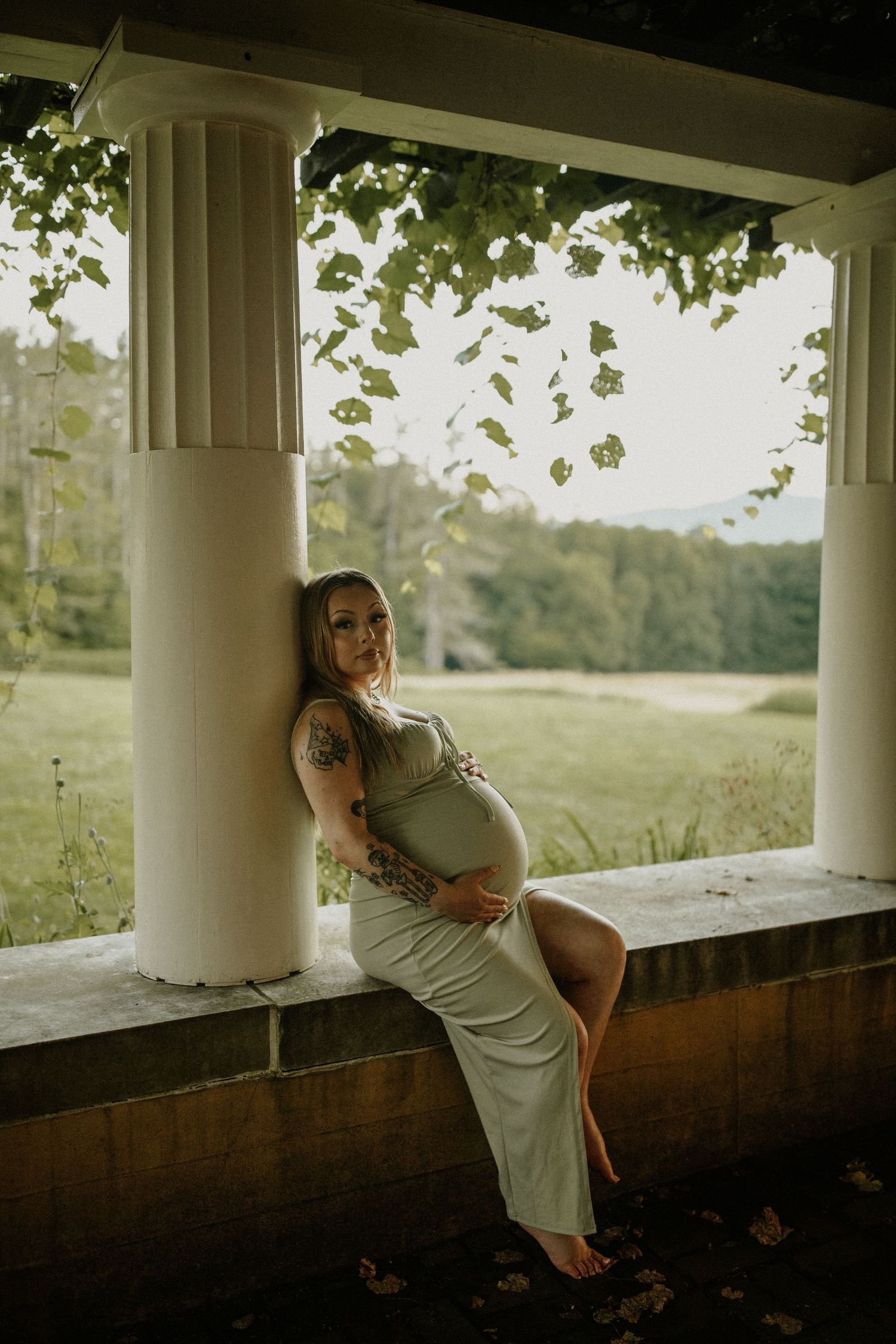 Maternity photo under a columned porch with green vines and golden afternoon light.