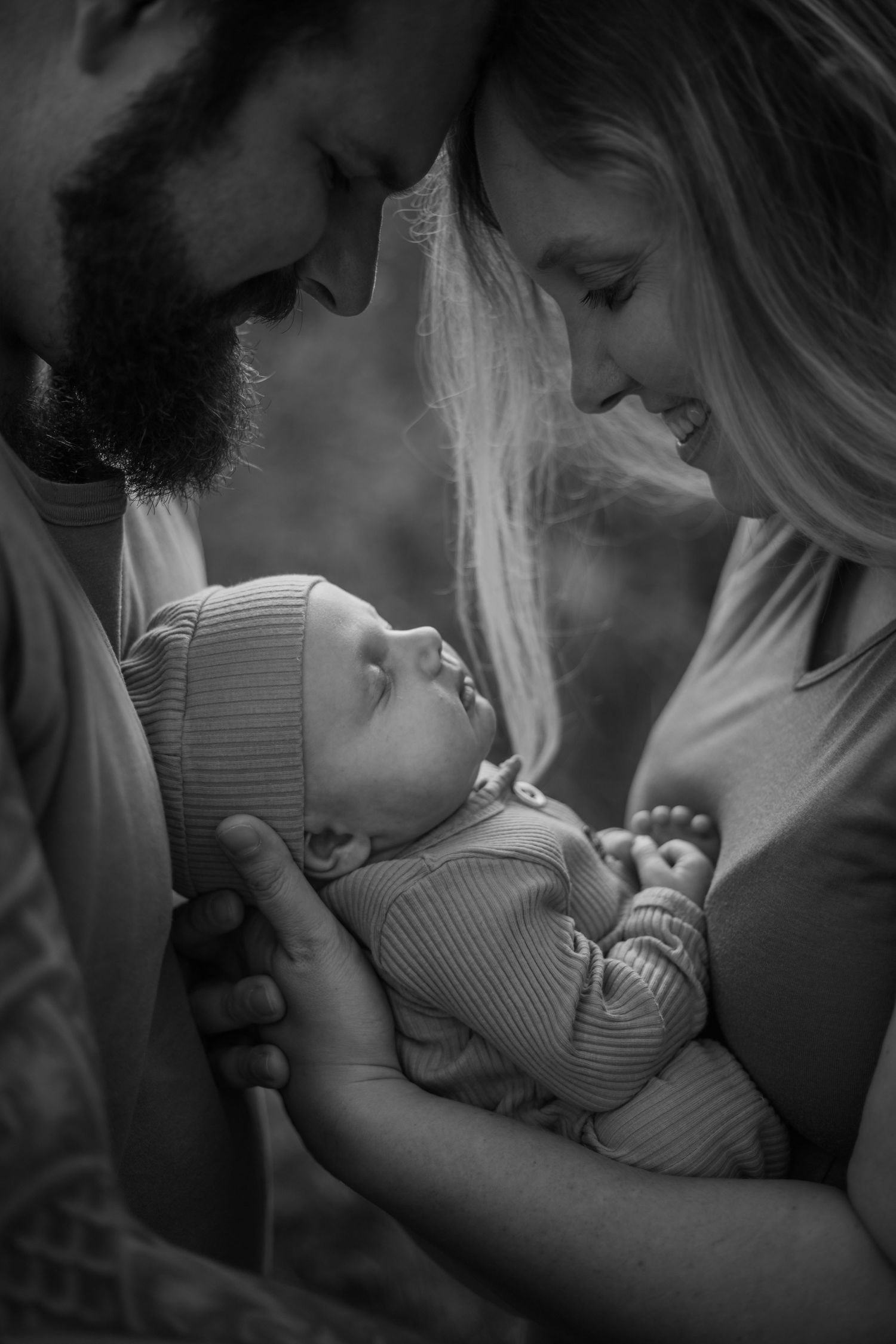 A tender black and white portrait of parents lovingly gazing at their newborn baby wearing a knit cap.