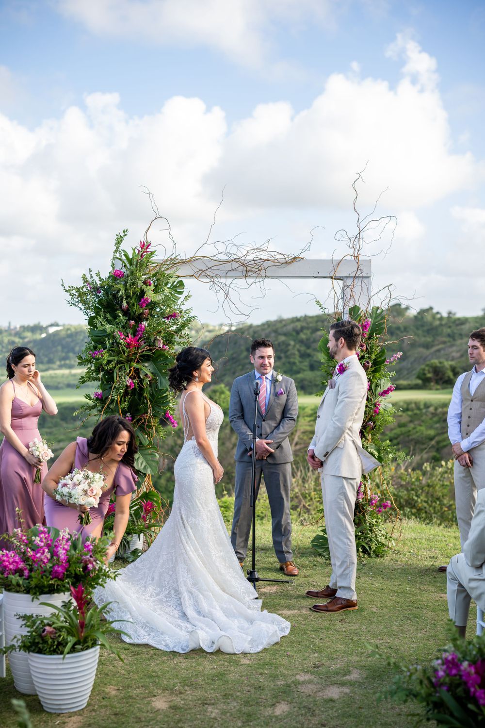 Outdoor wedding ceremony under a floral arch with pink blossoms and natural branches against a scenic hillside backdrop.