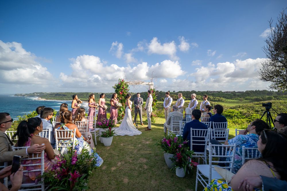 Destination wedding ceremony setup on a cliff overlooking the ocean with white chairs and tropical flower decorations.