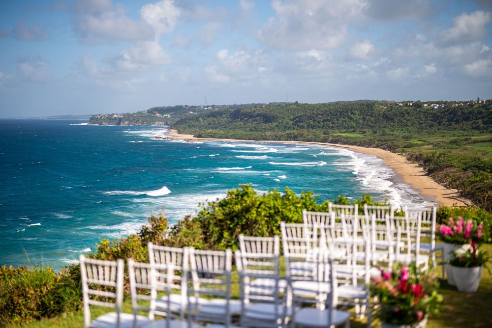White wedding chairs set up on a clifftop overlooking a beautiful turquoise ocean coastline.