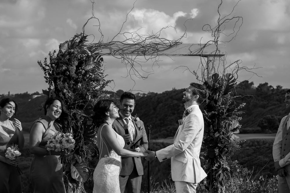Black and white photo of a wedding ceremony under a rustic branch arch with scenic views.