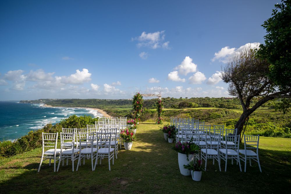 White wedding chairs set up for outdoor ceremony overlooking scenic coastal cliffs and blue ocean under sunny sky.