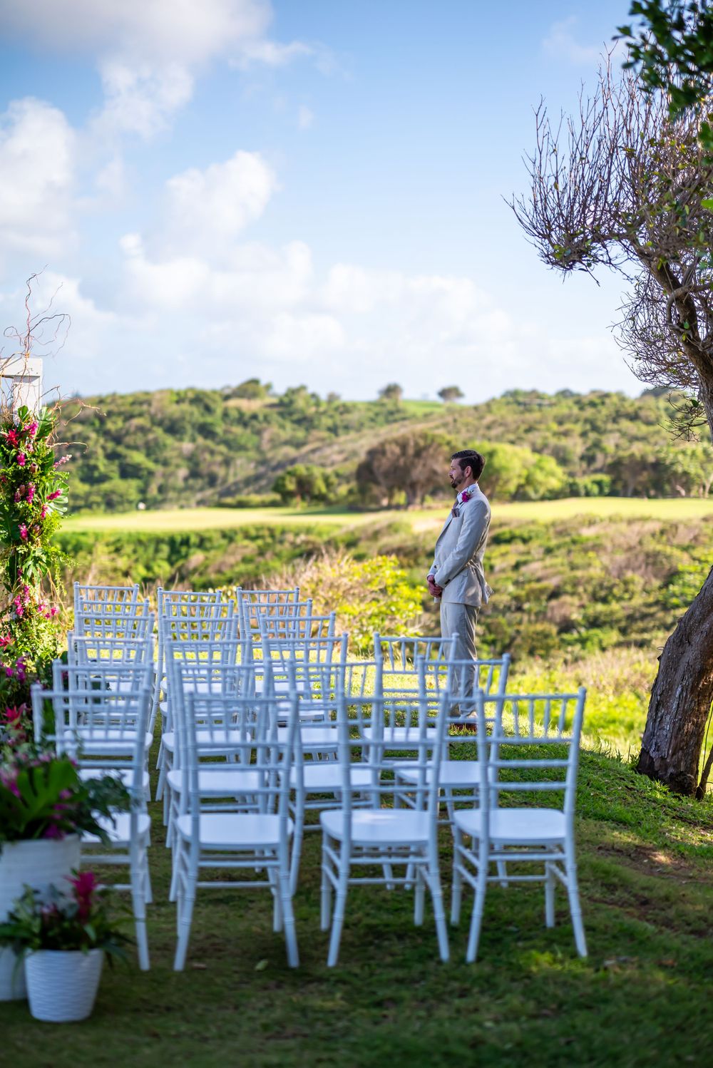 White ceremony chairs set up on grassy hillside with coastal views and person standing.