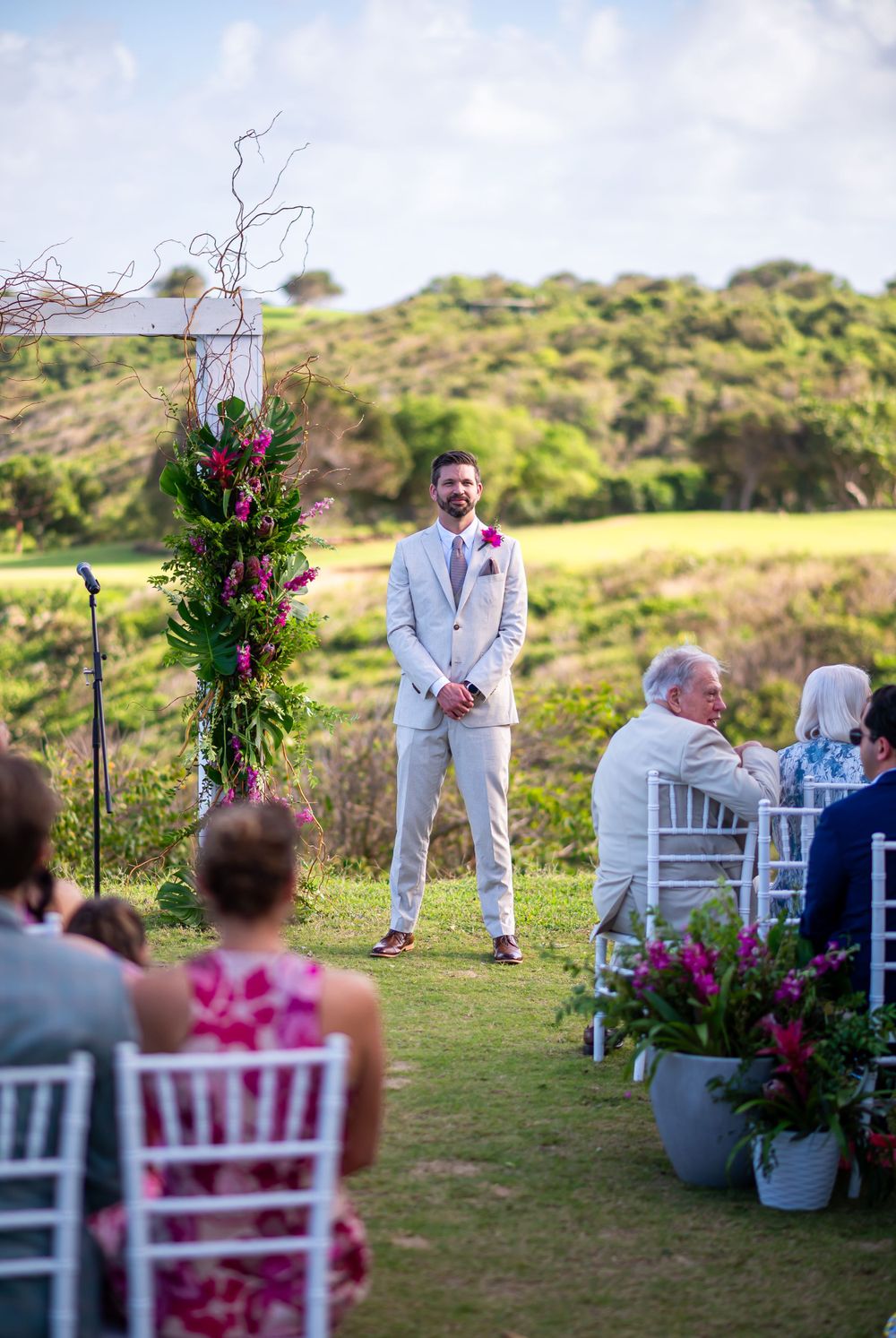 Wedding ceremony setup with floral arch and white chairs on coastal hilltop.