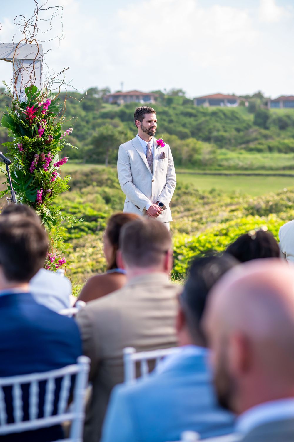 Outdoor wedding ceremony with guests seated on white chairs facing decorated floral arch.