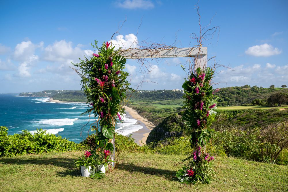 Tropical wedding ceremony setup with floral arch overlooking coastal cliffs and ocean views.