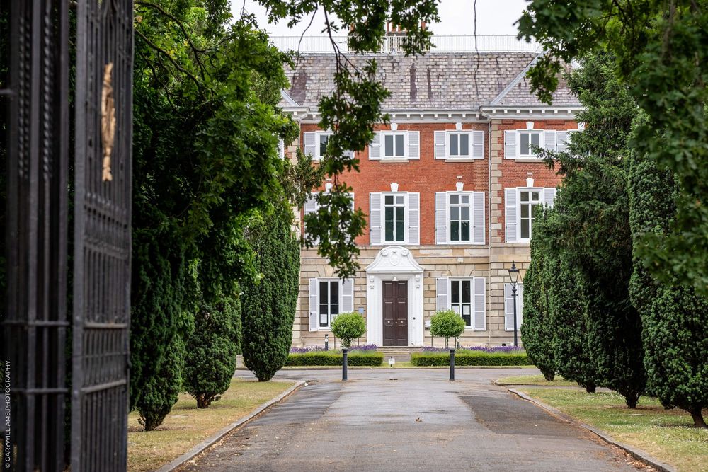 A grand red brick mansion with white trim and symmetrical windows viewed through a tree-lined driveway entrance.