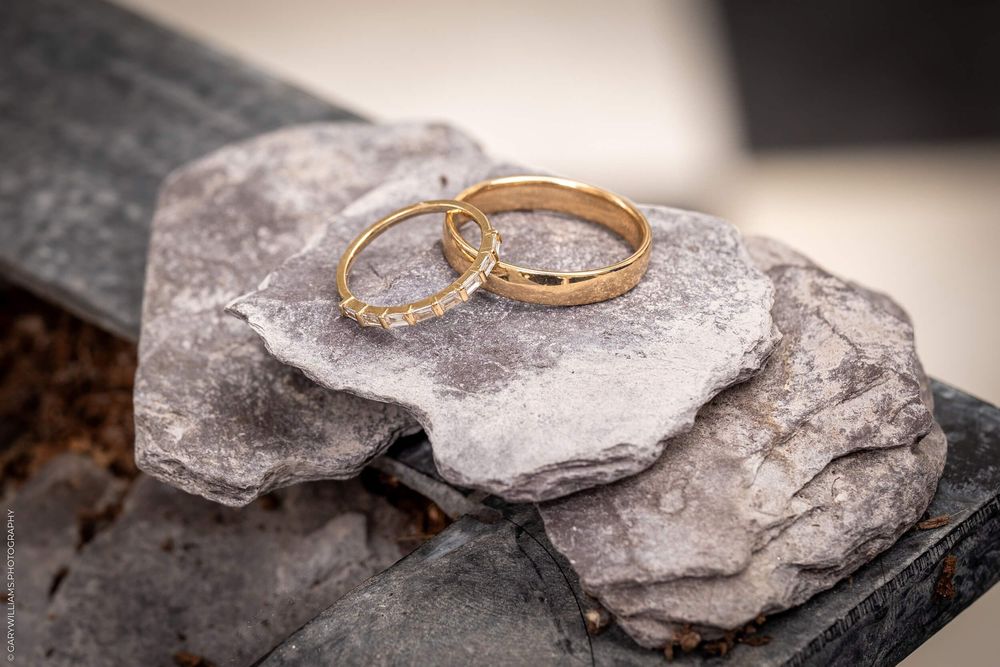 Two golden wedding bands with diamonds rest on a rough grey stone surface in soft lighting.