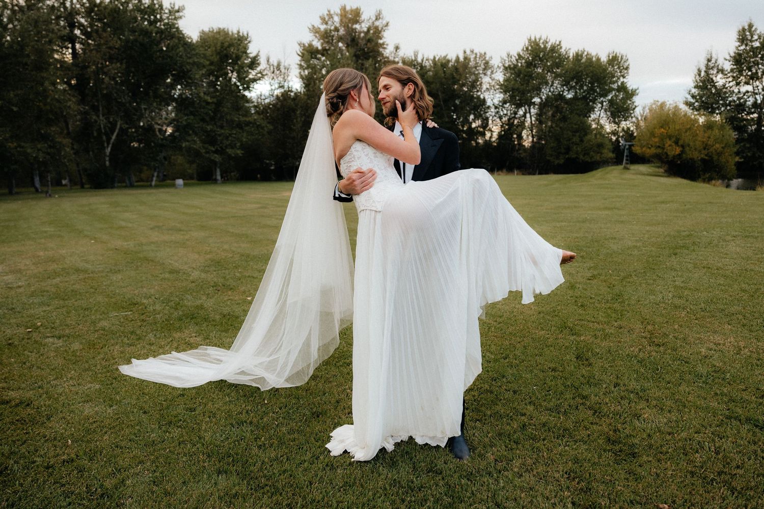 A newly married couple sharing an intimate moment outdoors as the groom carries the bride in her flowing white dress and veil.