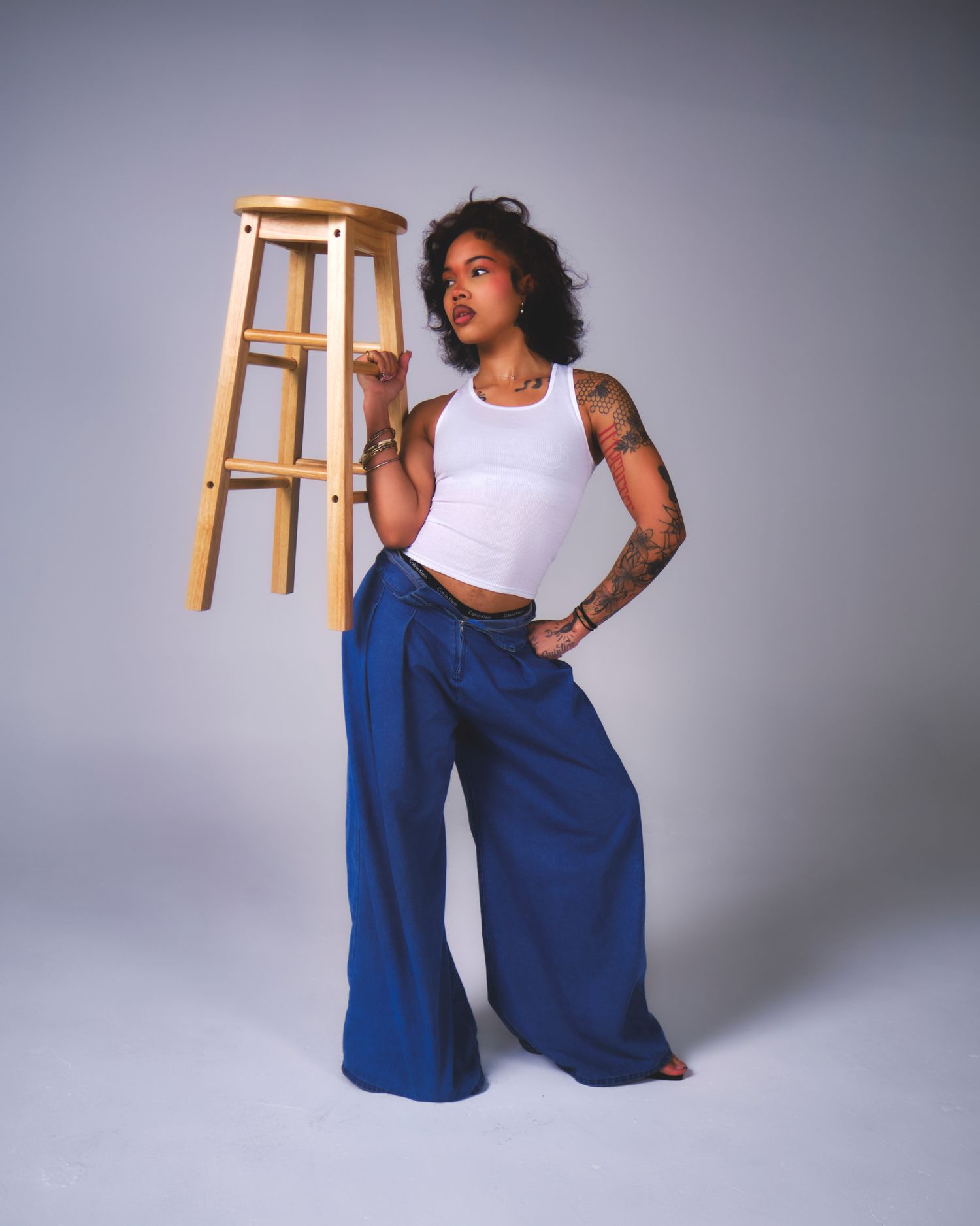 Fashion model in white crop top and wide-leg blue pants poses with wooden stool against gray backdrop.