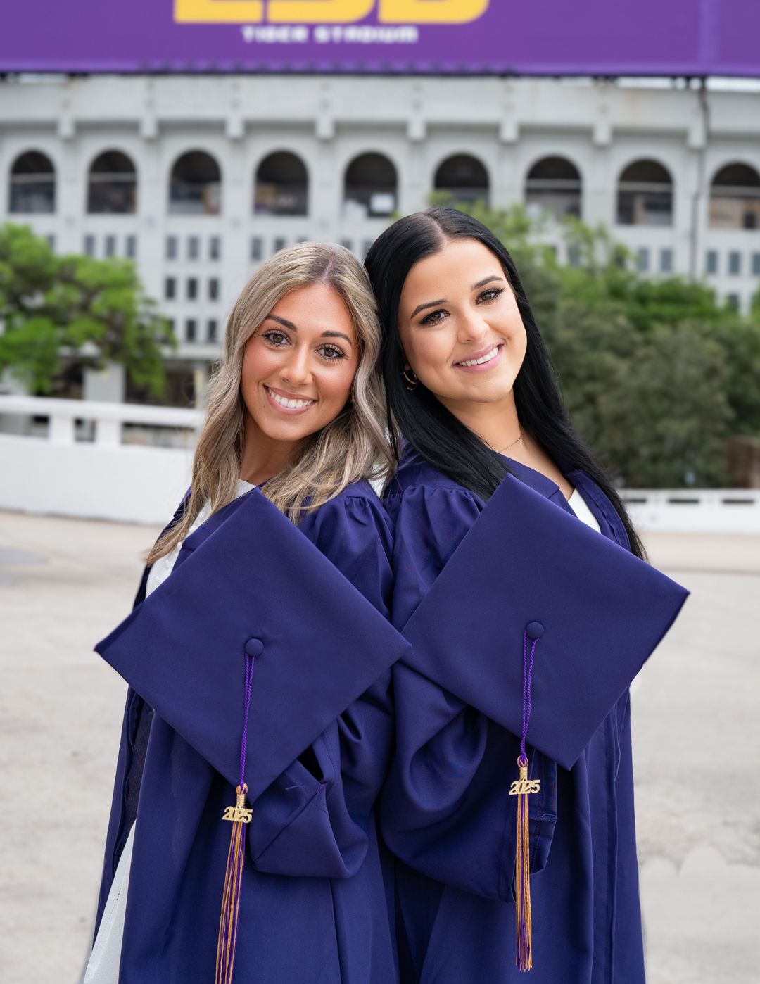 Two graduates in purple graduation gowns and caps posing together outdoors with LSU Stadium in background.