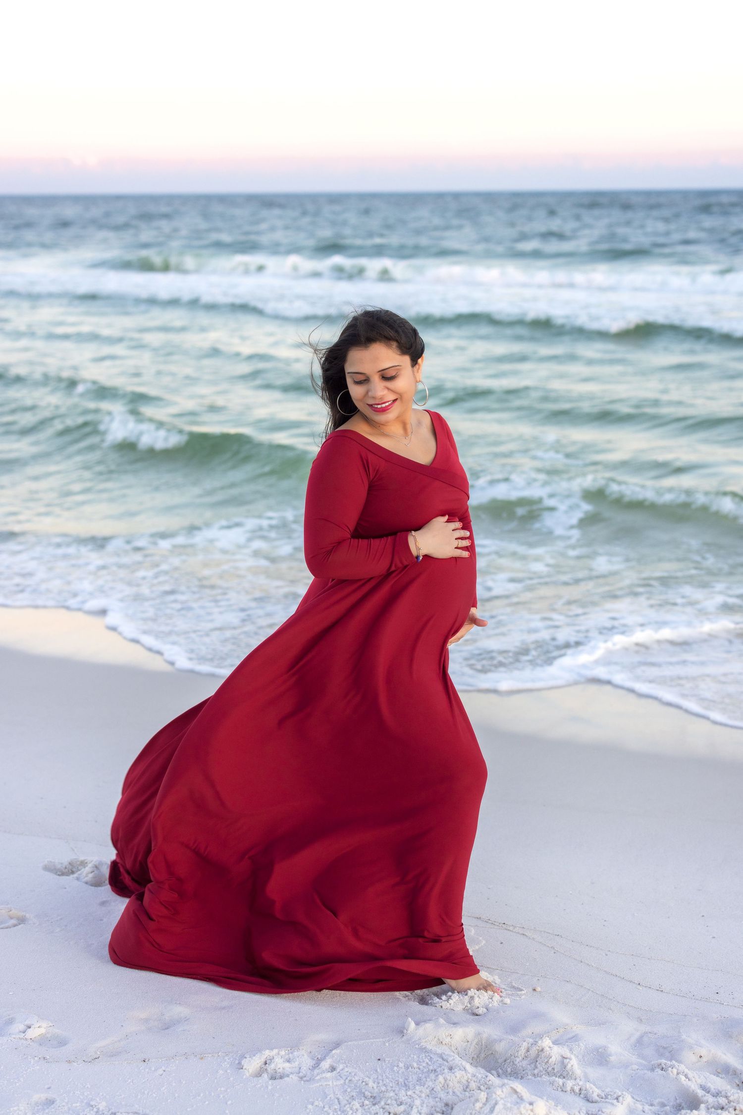 A flowing red maxi dress catches the breeze as someone poses gracefully on a beach at sunset.