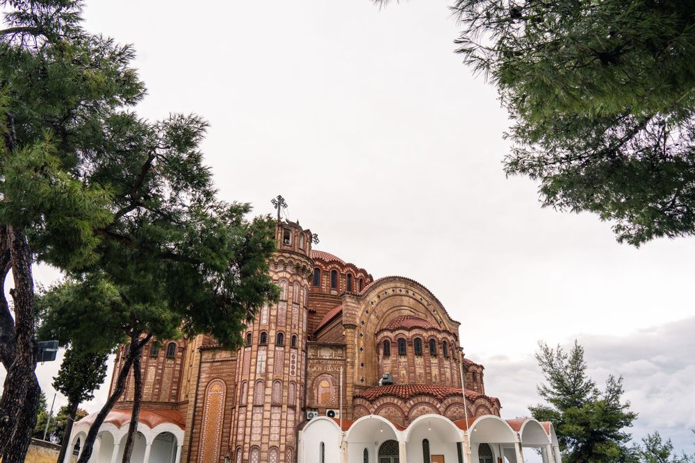 Byzantine-style church with red brick domes and ornate architecture viewed through green trees against cloudy sky.