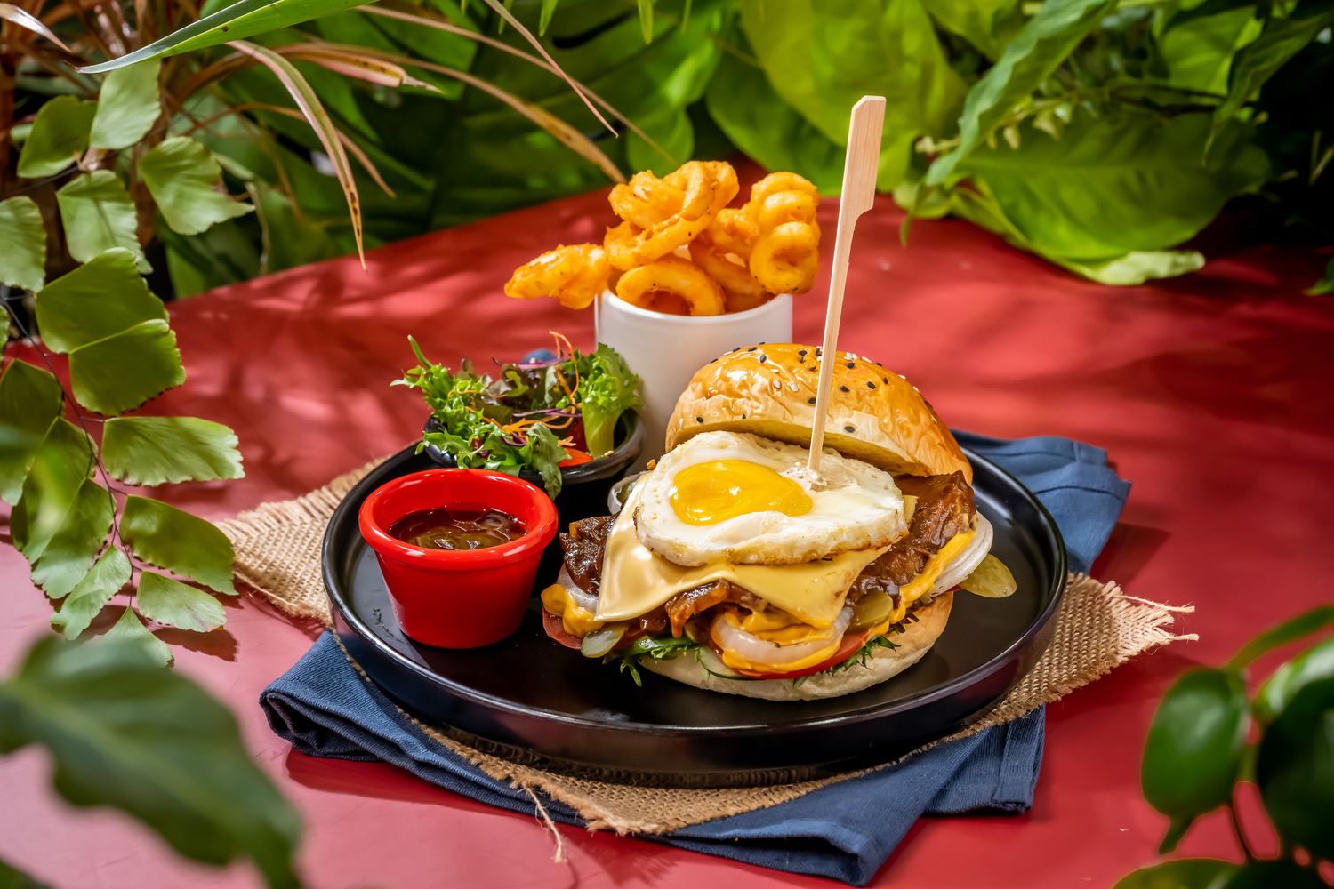 Gourmet burgers with fries and condiments served on red tablecloth.