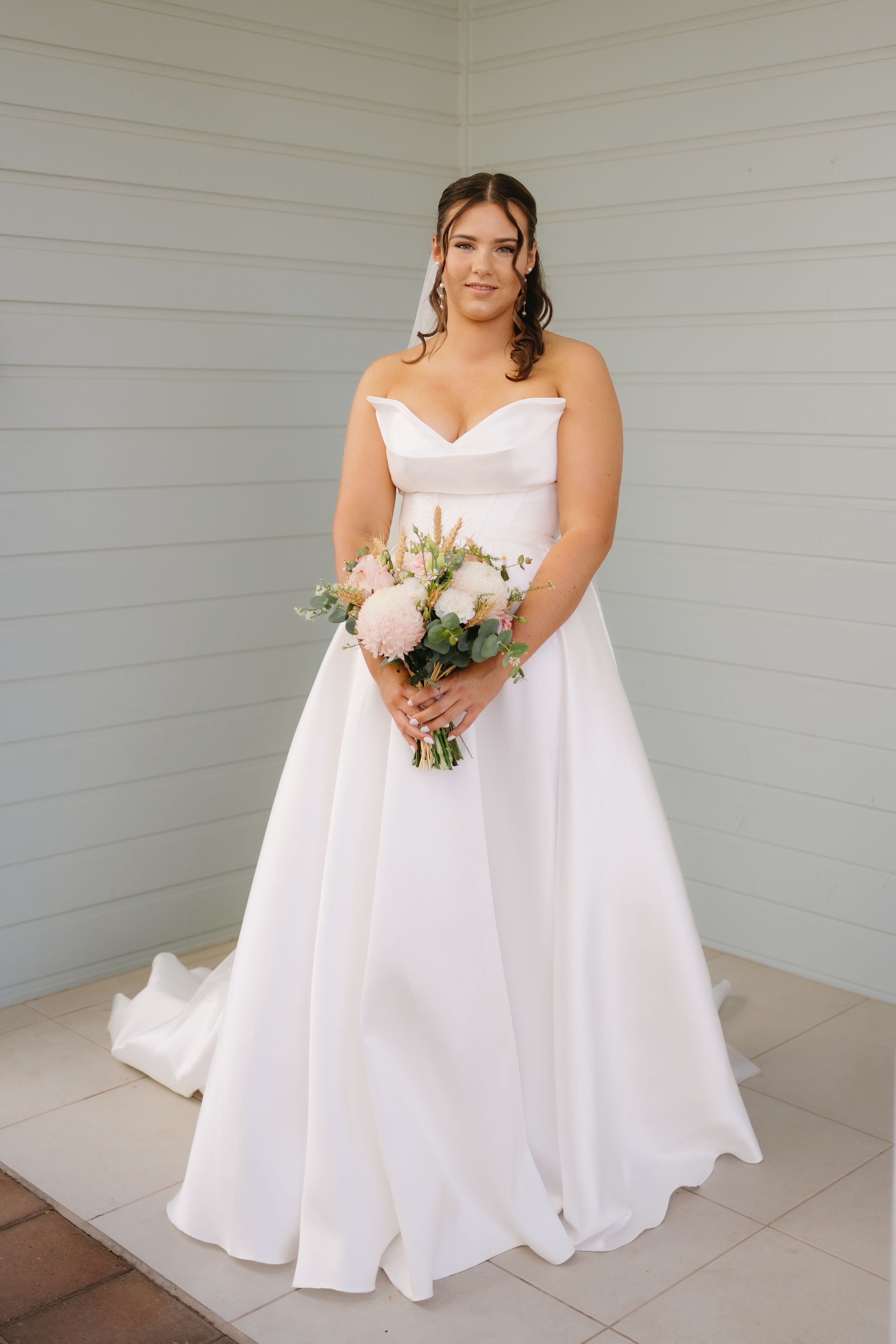 Bride in elegant white strapless wedding gown holds delicate bouquet against gray wall backdrop.