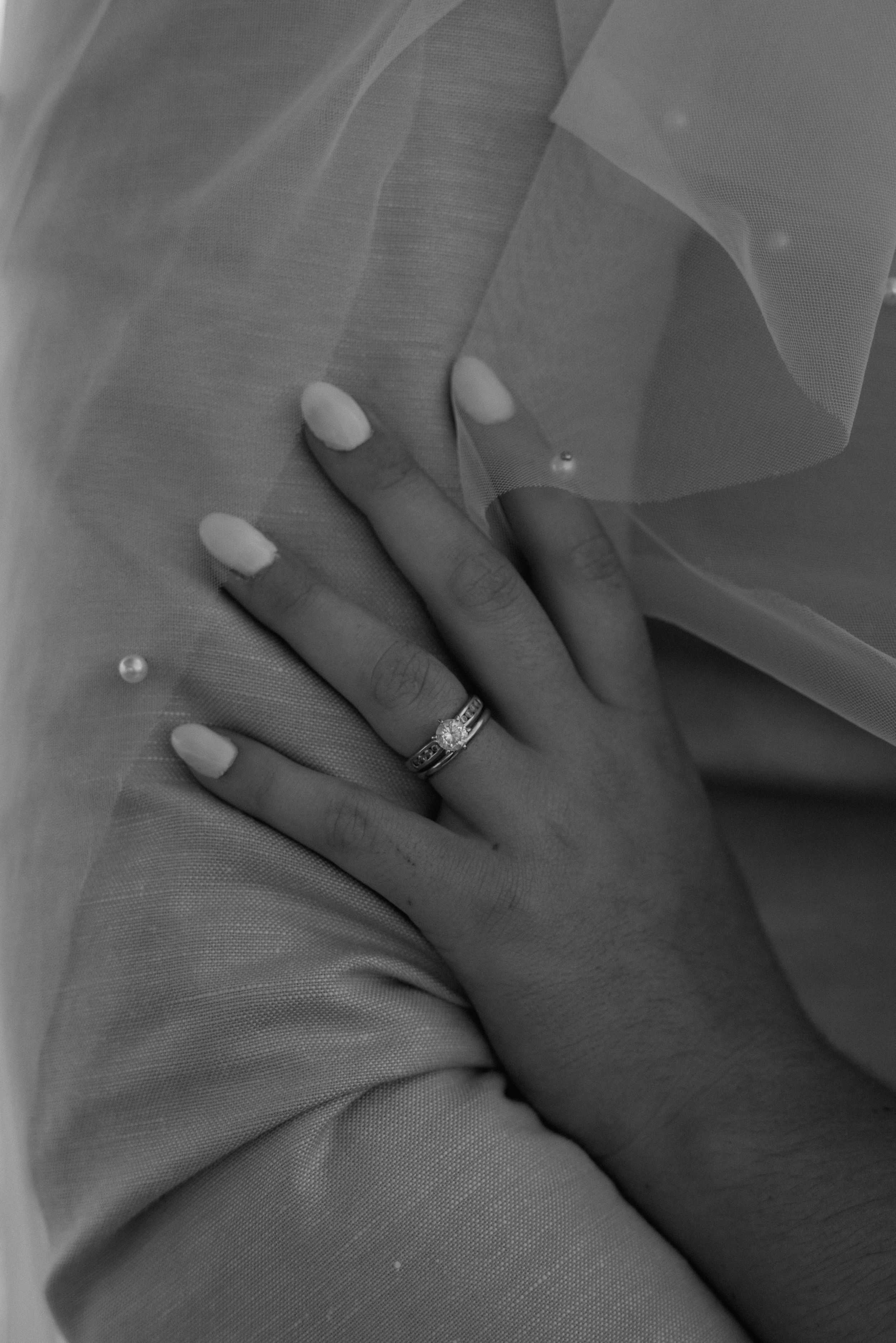 Black and white close-up of hand with wedding ring and manicured nails against tulle veil.