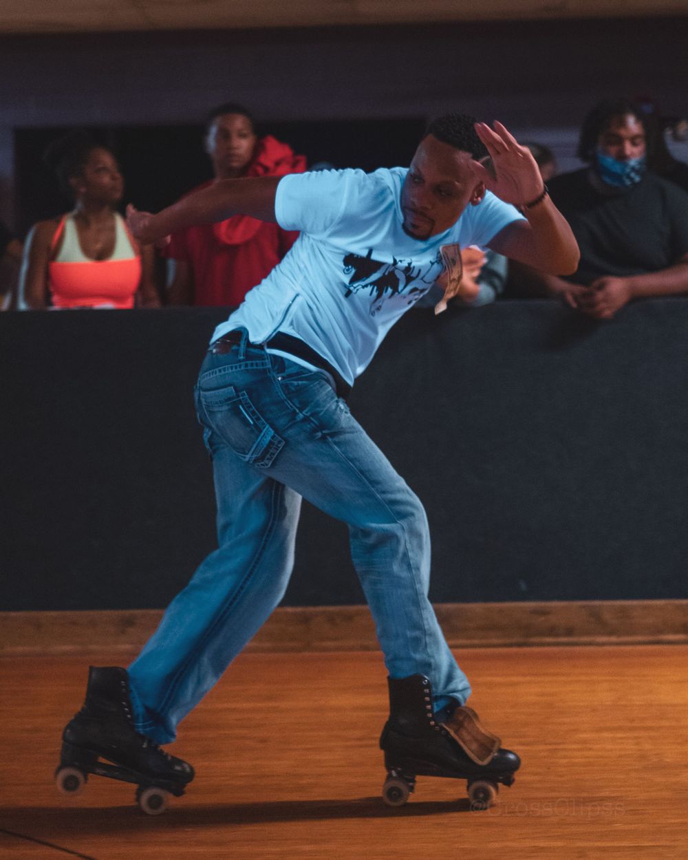 Roller skater in light blue shirt and jeans performs dynamic moves on wooden rink floor.