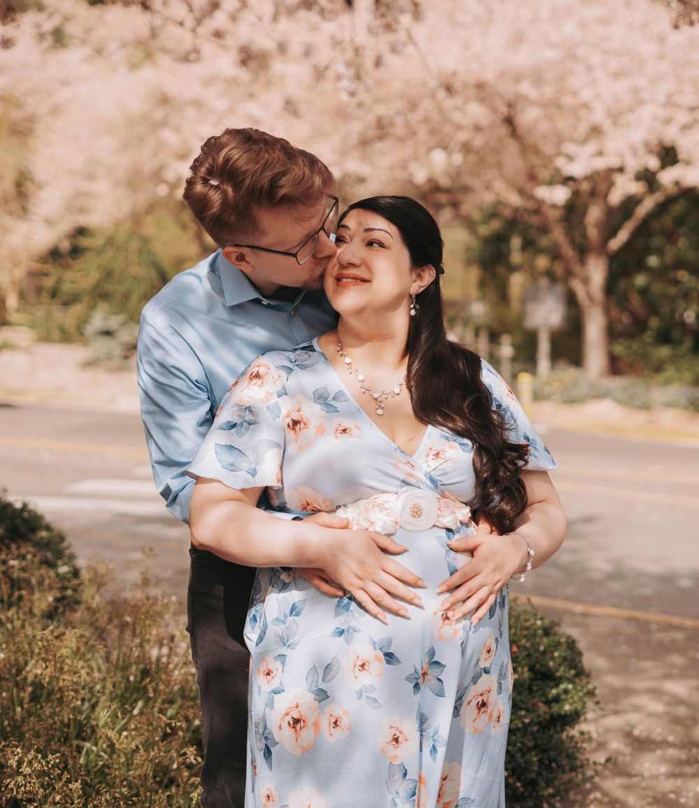 Couple embracing under blooming cherry blossom trees, man kissing womans cheek as she cradles baby bump.
